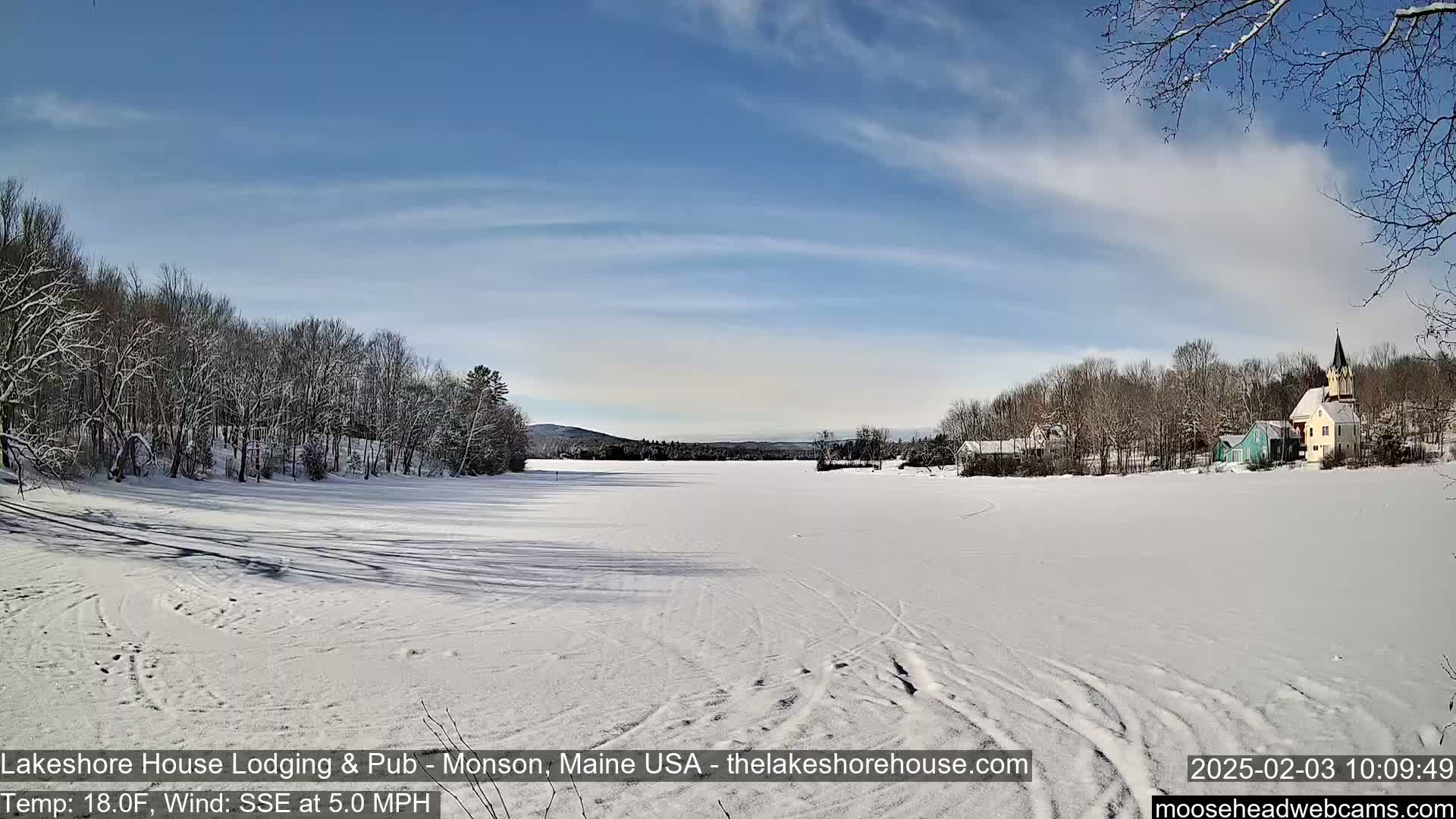 A snow-covered frozen lake stretches out before a line of snow-dusted trees and buildings under a mostly clear, bright blue sky.