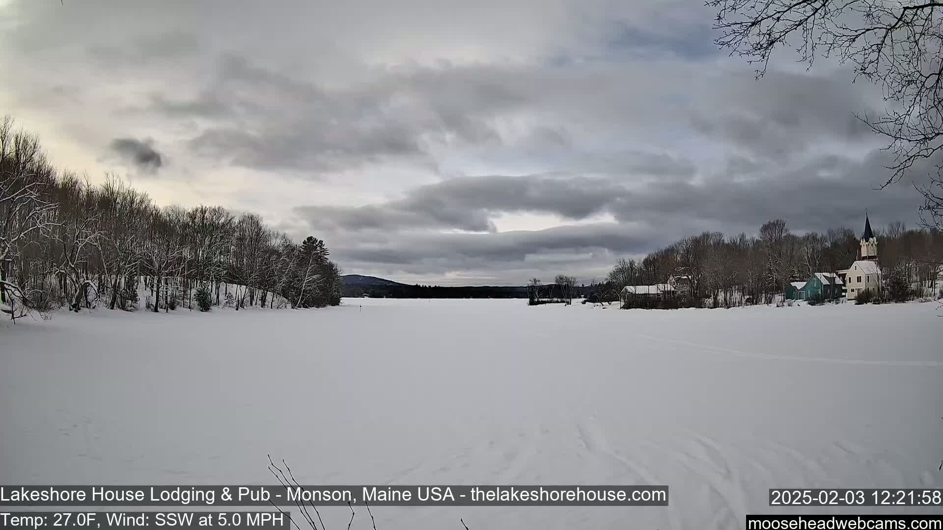 A snow-covered frozen lake stretches out before a village of snow-dusted buildings under a cloudy sky.