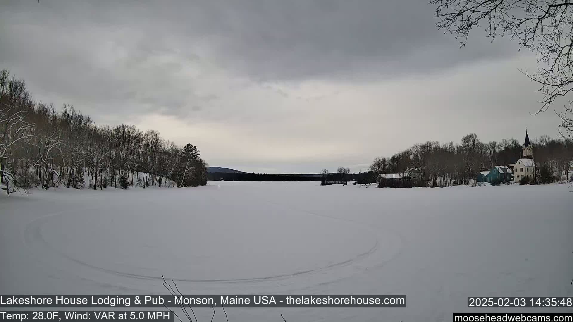 A snow-covered frozen lake is seen under an overcast sky, with snow-covered trees lining the shore and buildings visible in the distance.