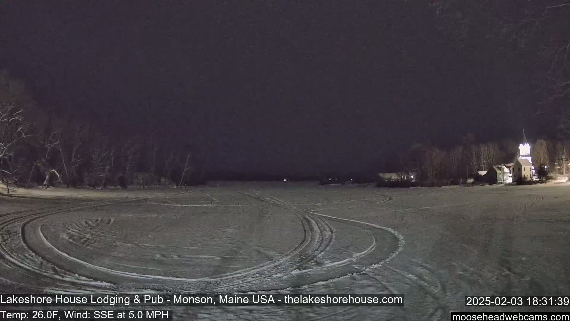 A snow-covered, flat expanse with tire tracks under a dark night sky shows a few dimly lit buildings in the distance.