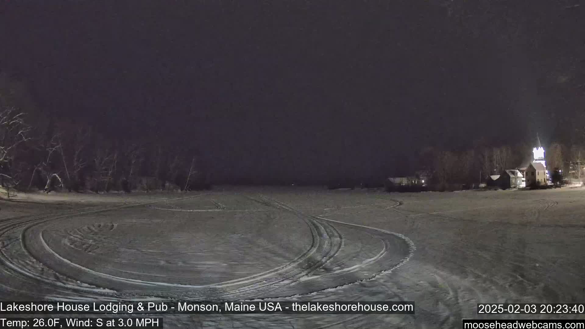 A snow-covered field at night, with tire tracks curving across its surface, and buildings faintly visible in the distance under a dark sky.