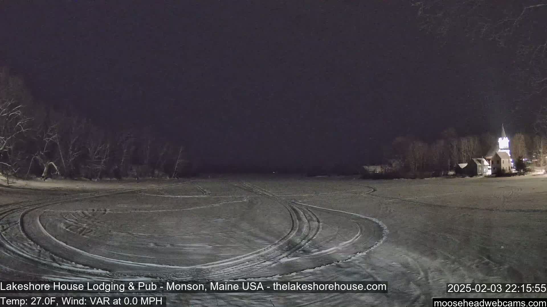 A snow-covered, flat expanse with tire tracks is illuminated at night, showing a building with a steeple in the distance under a dark sky.
