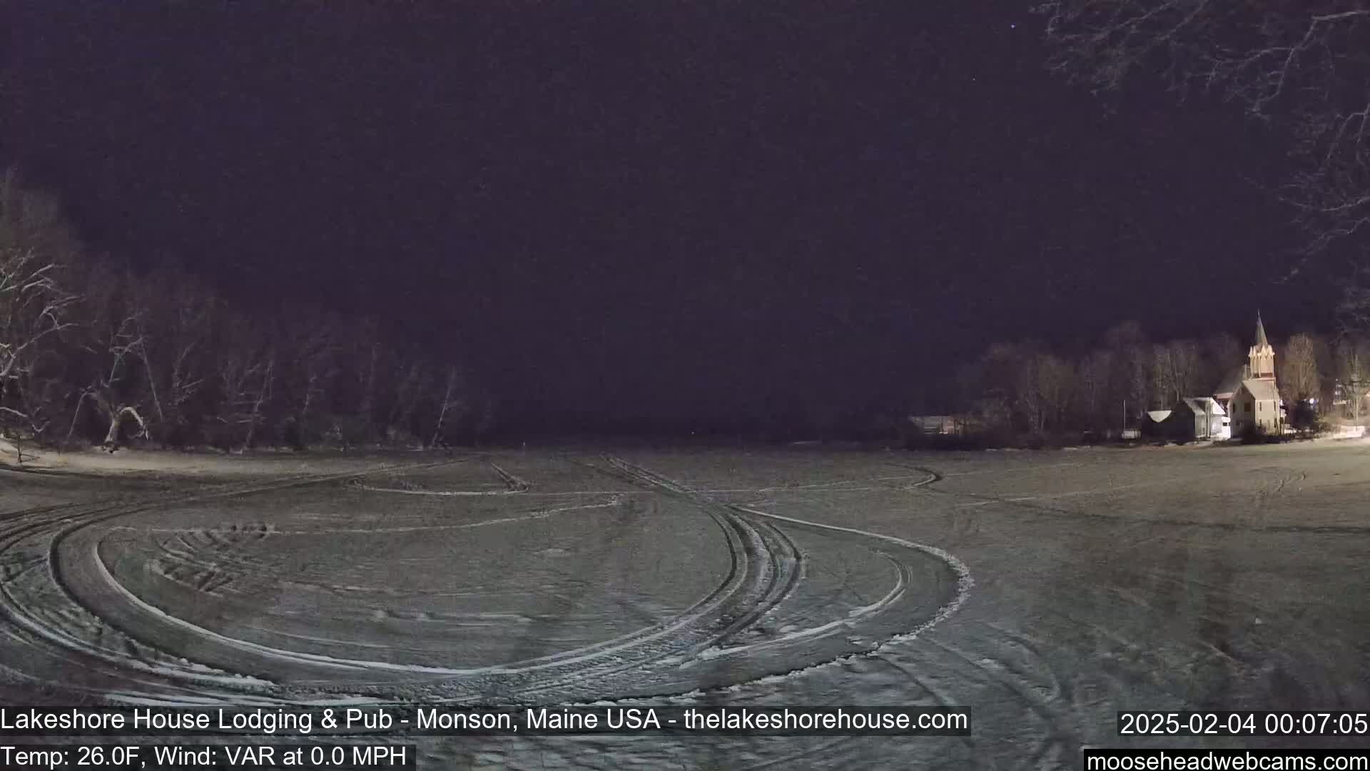 A snow-covered field with tire tracks shows a church and buildings in the distance under a dark night sky.