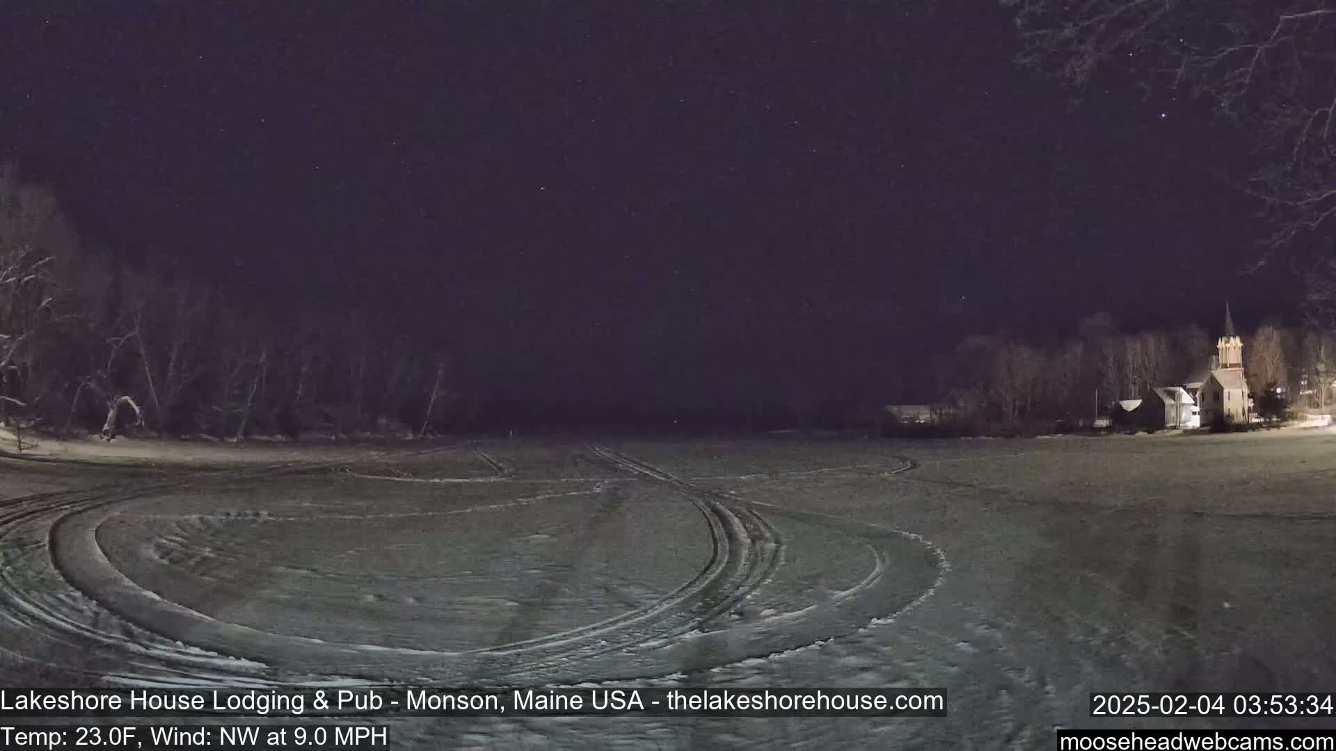 Under a clear, starry night sky, tire tracks crisscross a snow-covered field, with a church visible in the distance.
