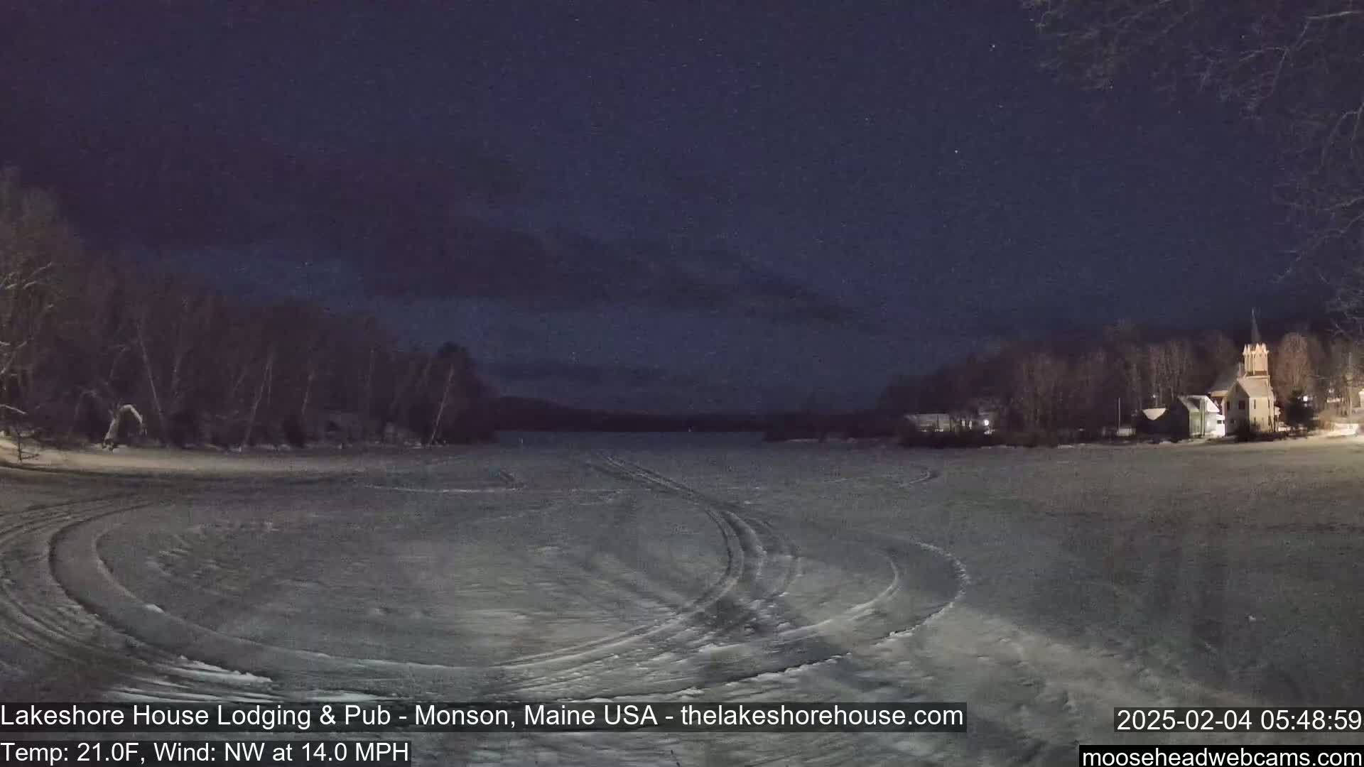 A snow-covered, frozen lake at night, with tire tracks visible on the ice and buildings in the distance under a dark, starry sky.