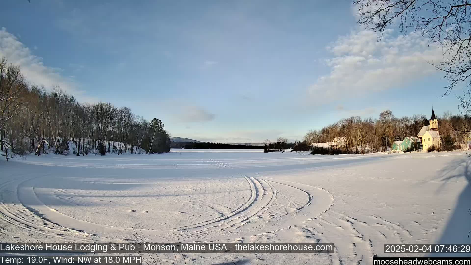 A frozen lake with tire tracks, surrounded by snow-covered trees and buildings under a mostly clear, sunny sky.