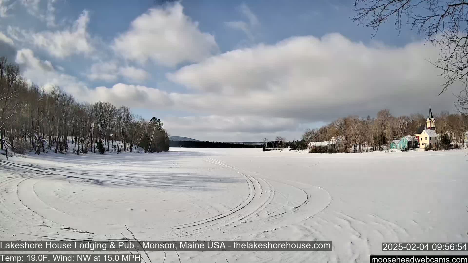 A snow-covered frozen lake with tire tracks is seen, with bare trees and a small village in the background under a partly cloudy sky.