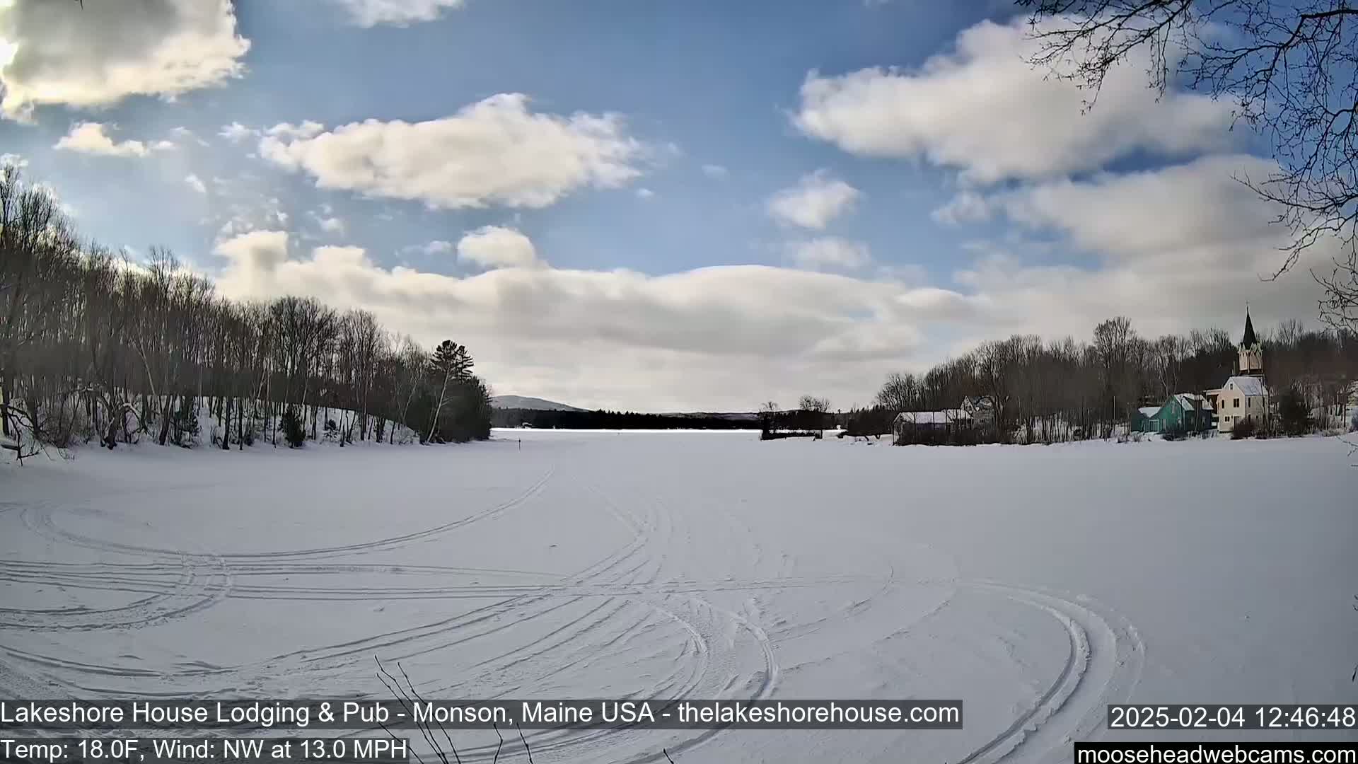 A frozen lake with tire tracks is seen under a partly cloudy sky, with buildings and trees visible on the far shore.