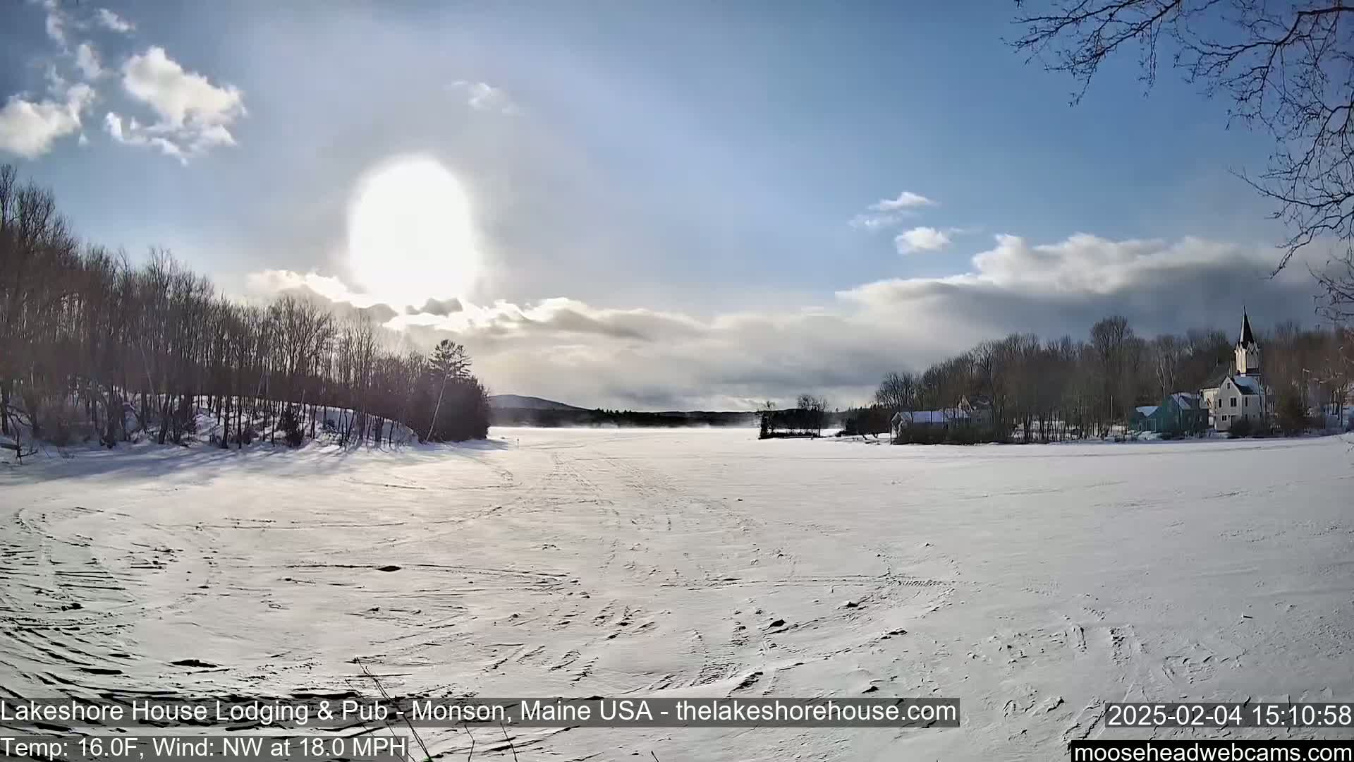 A snow-covered frozen lake stretches out under a partly cloudy, sunny sky, with bare trees lining the shore and a village visible in the distance.