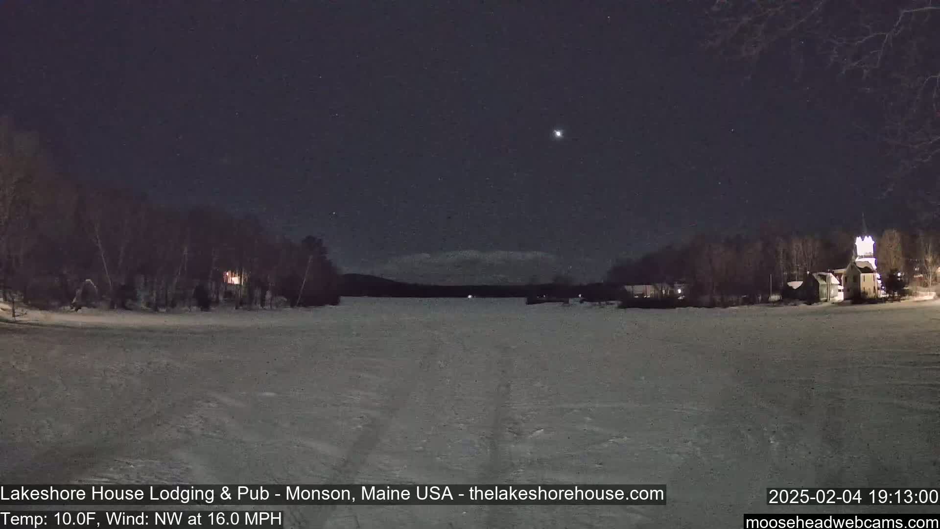 A snow-covered frozen lake is seen at night under a clear, starry sky, with a few lights visible on the far shore and trees lining the water's edge.