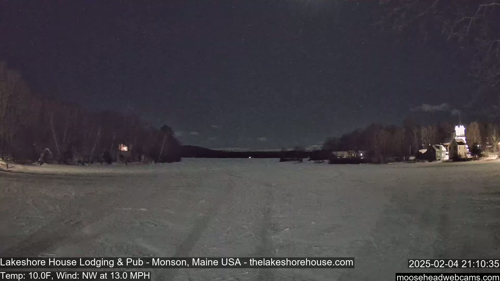 A snow-covered frozen lake is visible at night under a starry sky, with buildings and trees lining the shoreline.