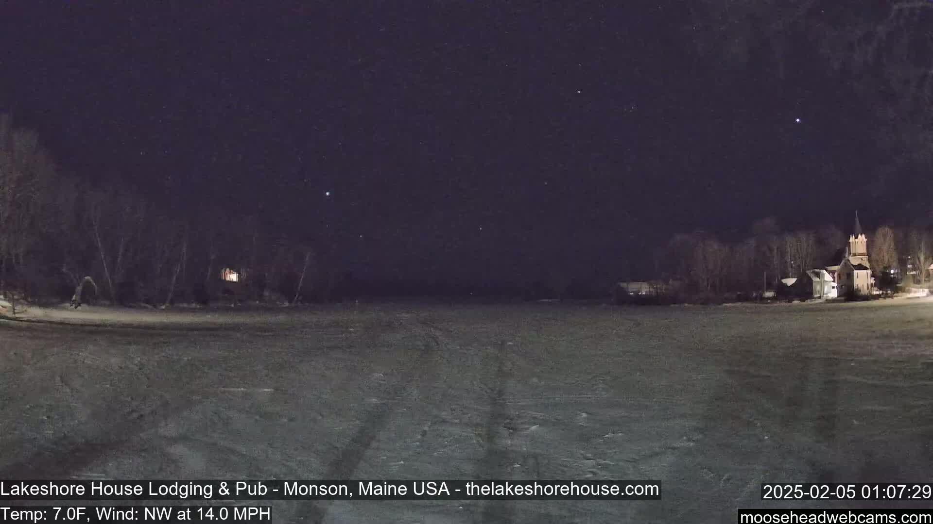 A snow-covered, dark expanse of land and frozen water stretches out under a clear, starlit night sky, with a few buildings and trees visible in the distance.