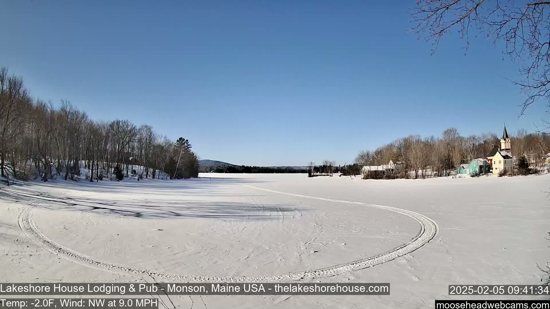 A frozen lake with tire tracks curves around a snow-covered shoreline, featuring a small village with colorful buildings under a clear blue sky on a cold, windy day.