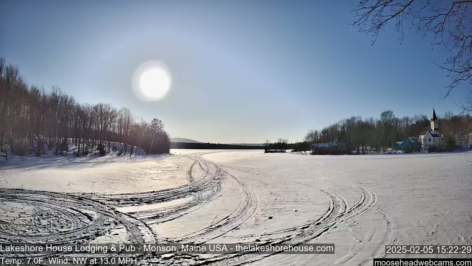 Tire tracks crisscross a snow-covered frozen lake under a bright, sunny sky, with a village and trees visible in the distance.