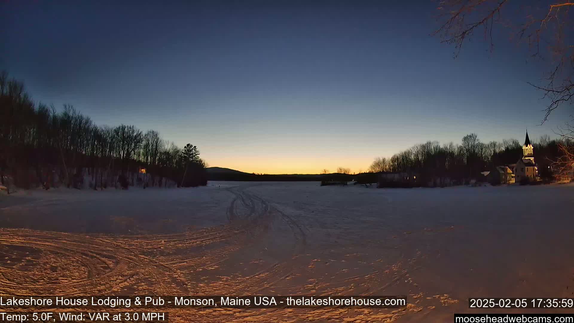 A frozen lake at twilight is partially illuminated by a setting sun, with a village and trees lining the shore in the background under a clear sky.