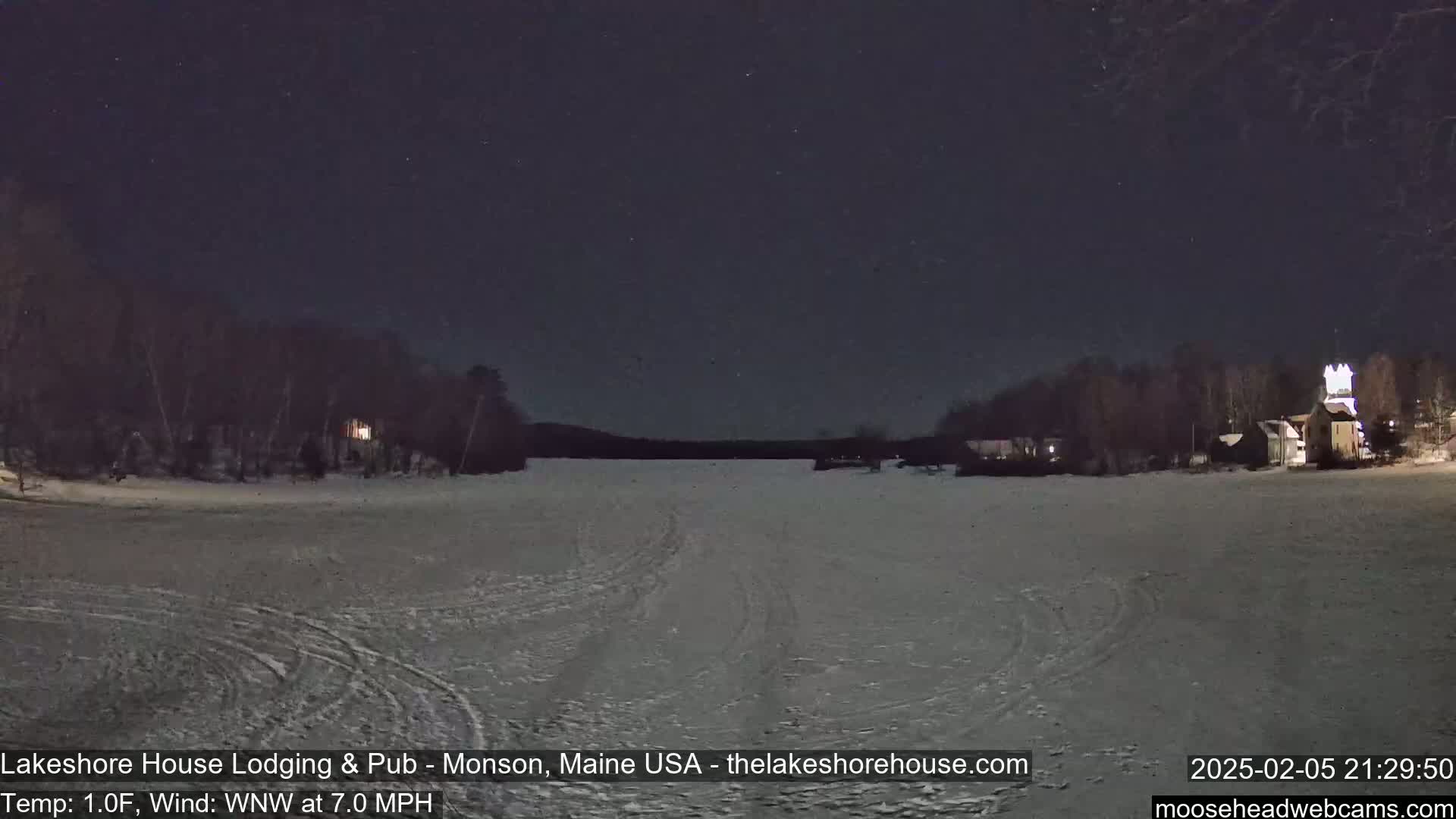 A snow-covered, frozen lake is seen at night under a starry sky, with buildings and trees visible on either shore.