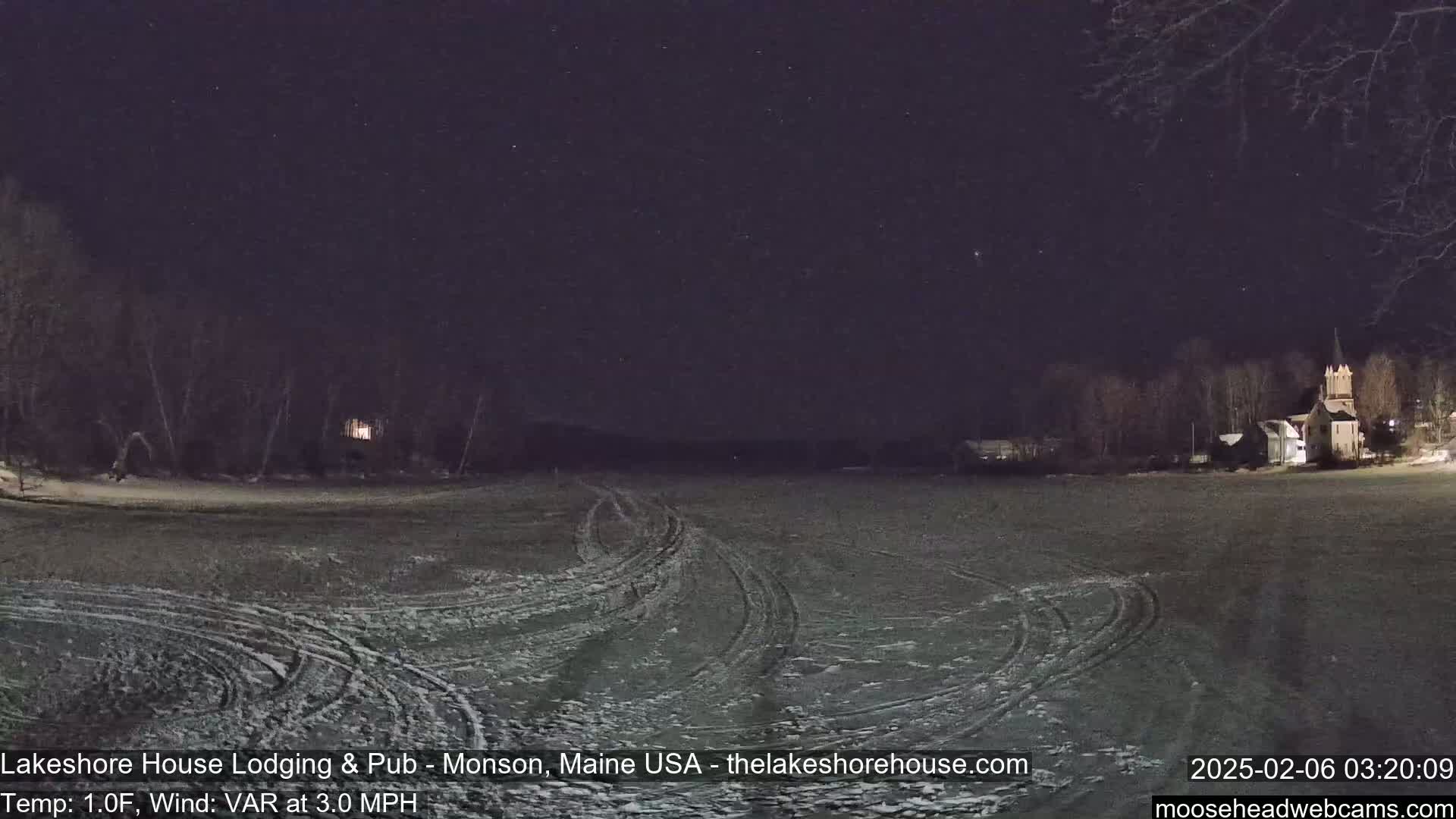 A snow-covered field with tire tracks is visible under a clear, starry night sky, with buildings and trees in the distance.