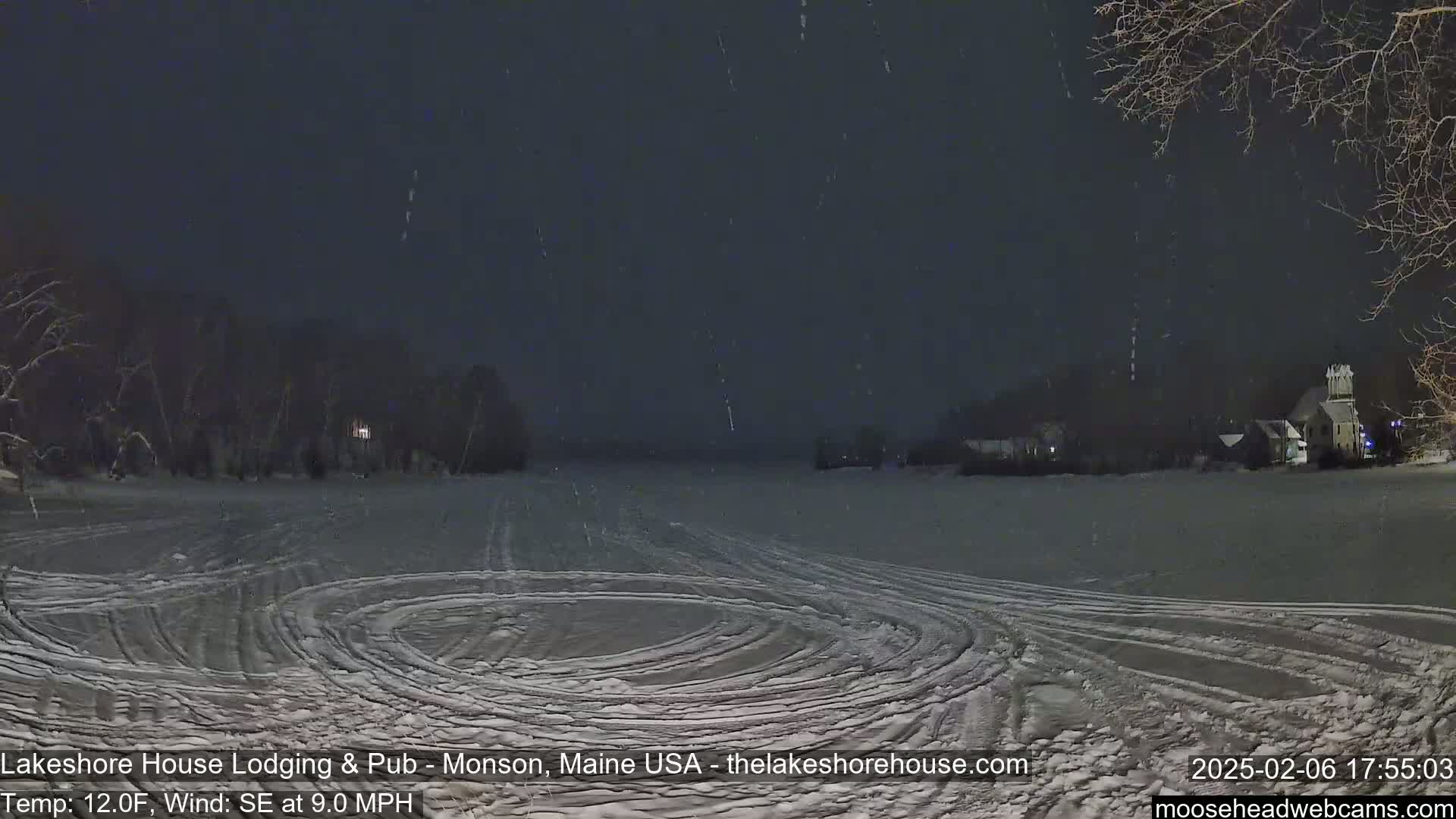 A snow-covered, flat expanse with tire tracks shows a nighttime scene of light snowfall, with buildings and trees faintly visible in the distance under a dark sky.