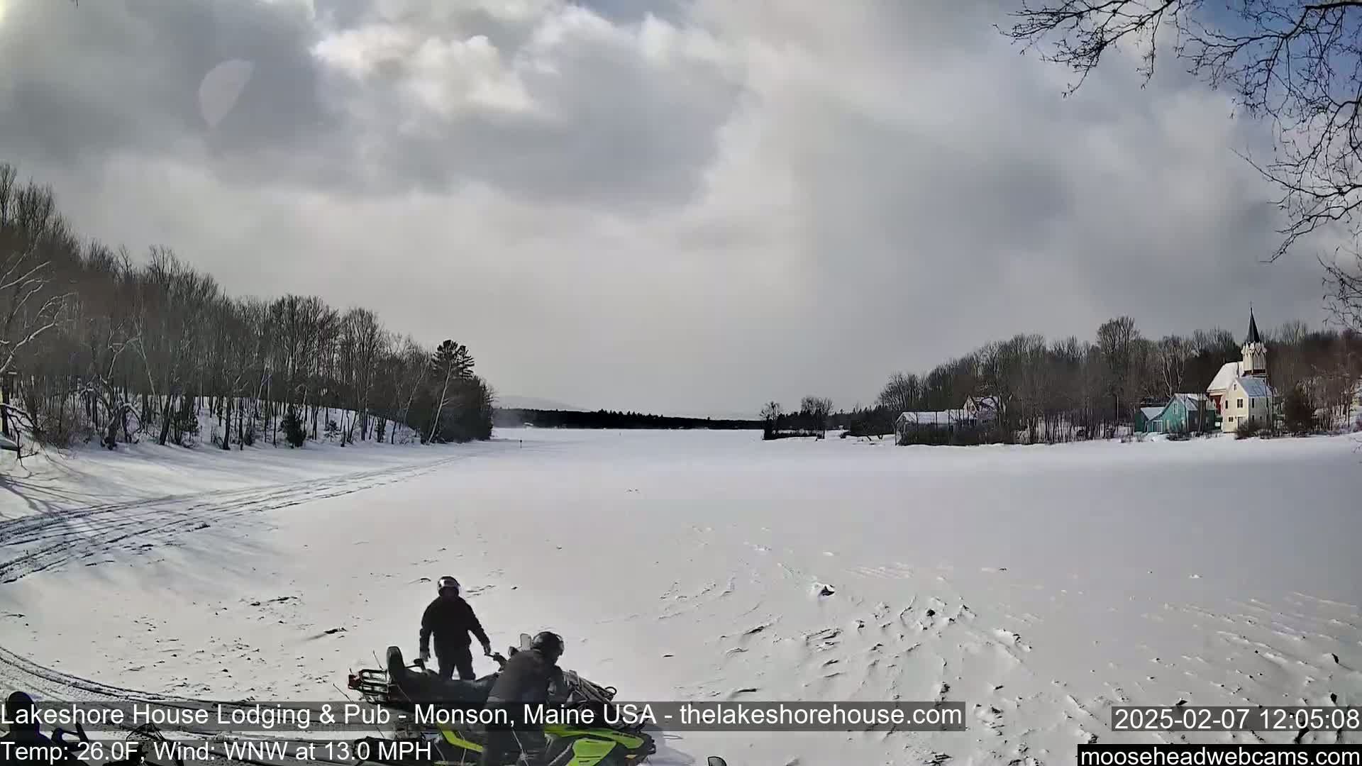 A snowmobile with two riders is on a frozen lake under a cloudy sky, with a village visible in the distance.
