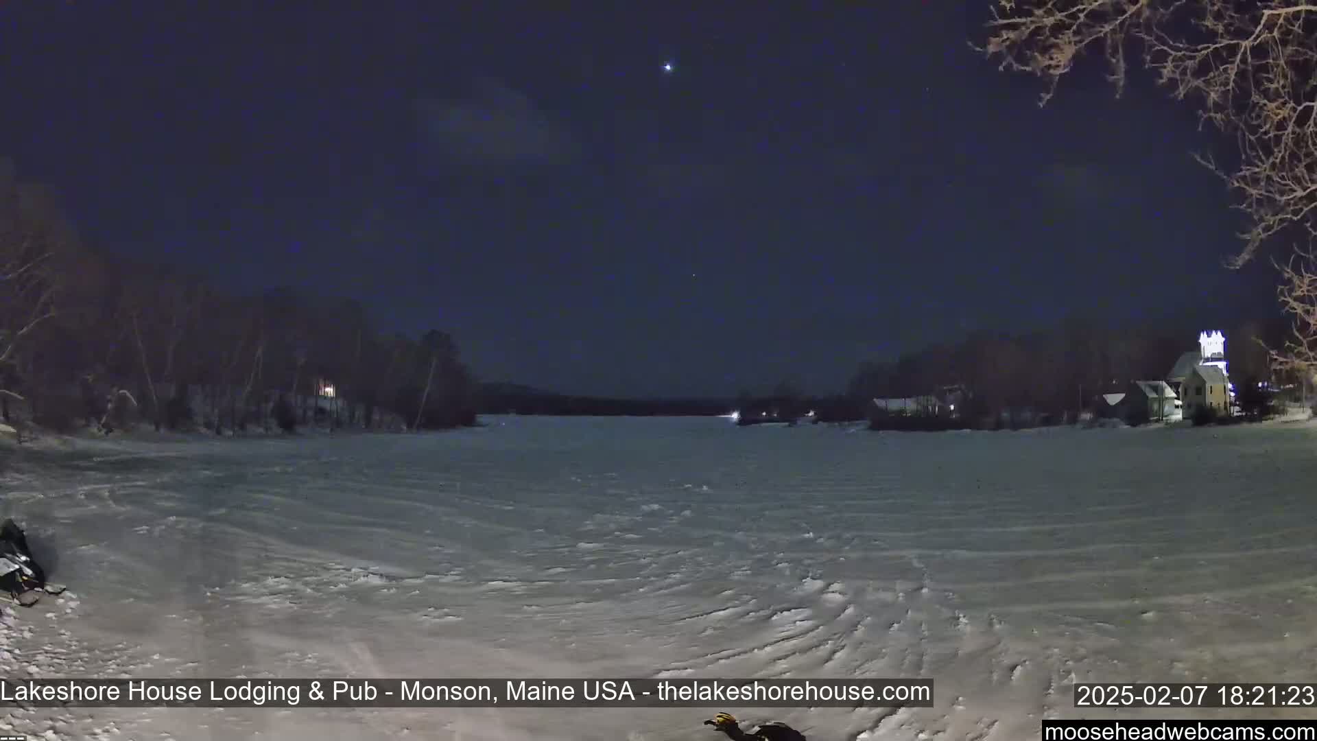 A snow-covered frozen lake is seen at night under a clear, dark sky with a few stars visible, with buildings and trees visible on the far shore.