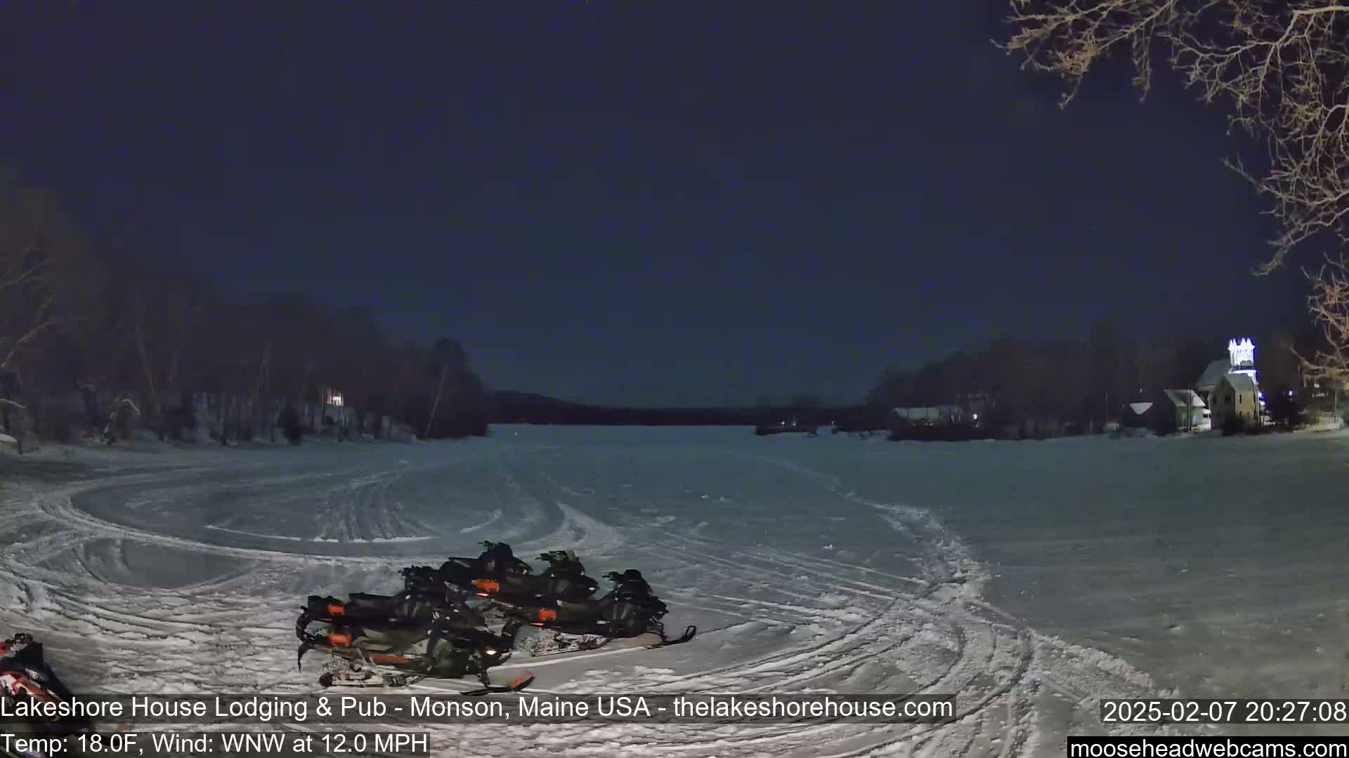 Several snowmobiles are parked on a snow-covered frozen lake at night under a dark sky, with a lightly lit town visible in the distance, and a 12 mph wind.
