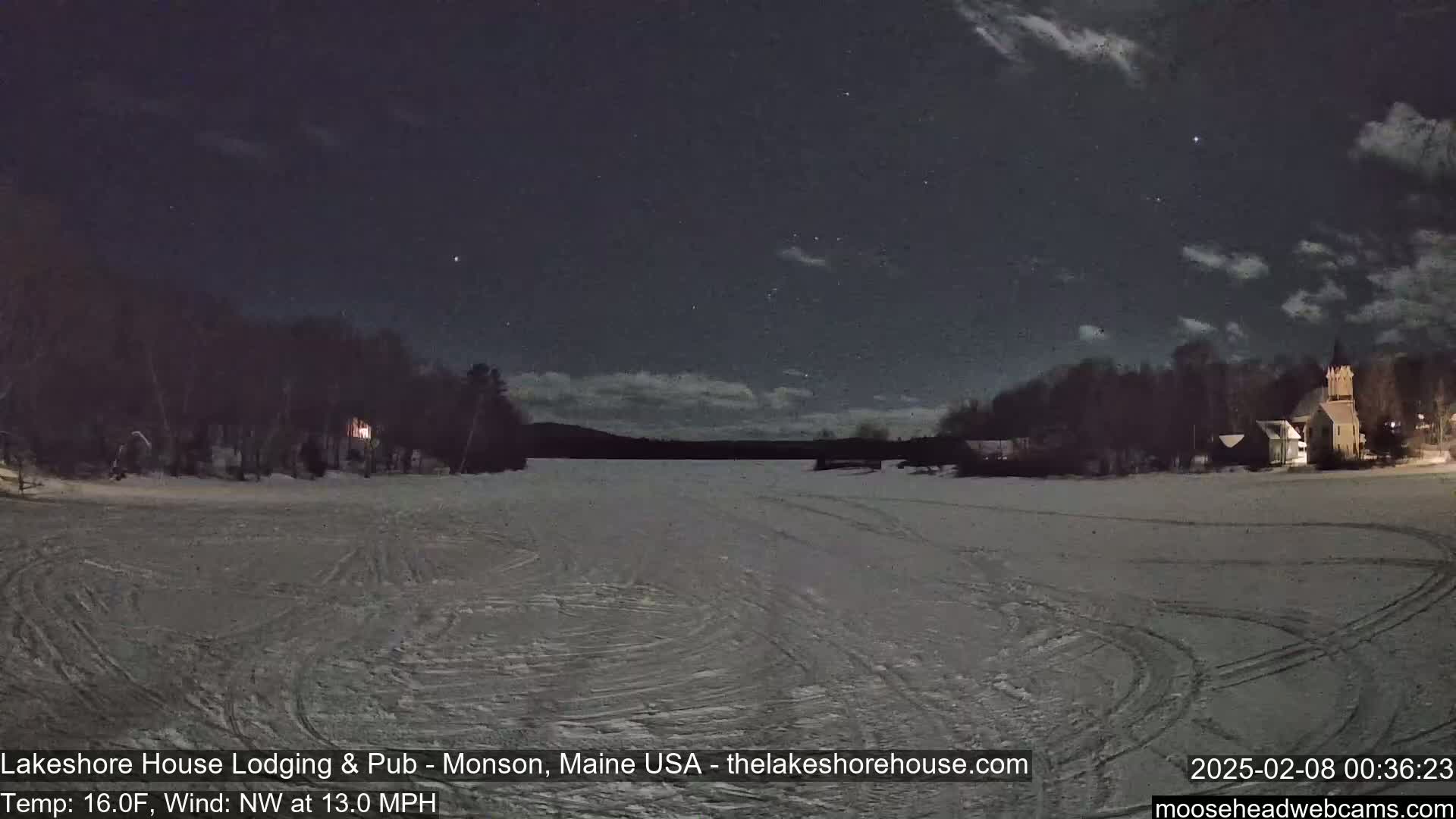 A snow-covered, frozen lake is visible under a partly cloudy night sky, with buildings and trees lining the shore.