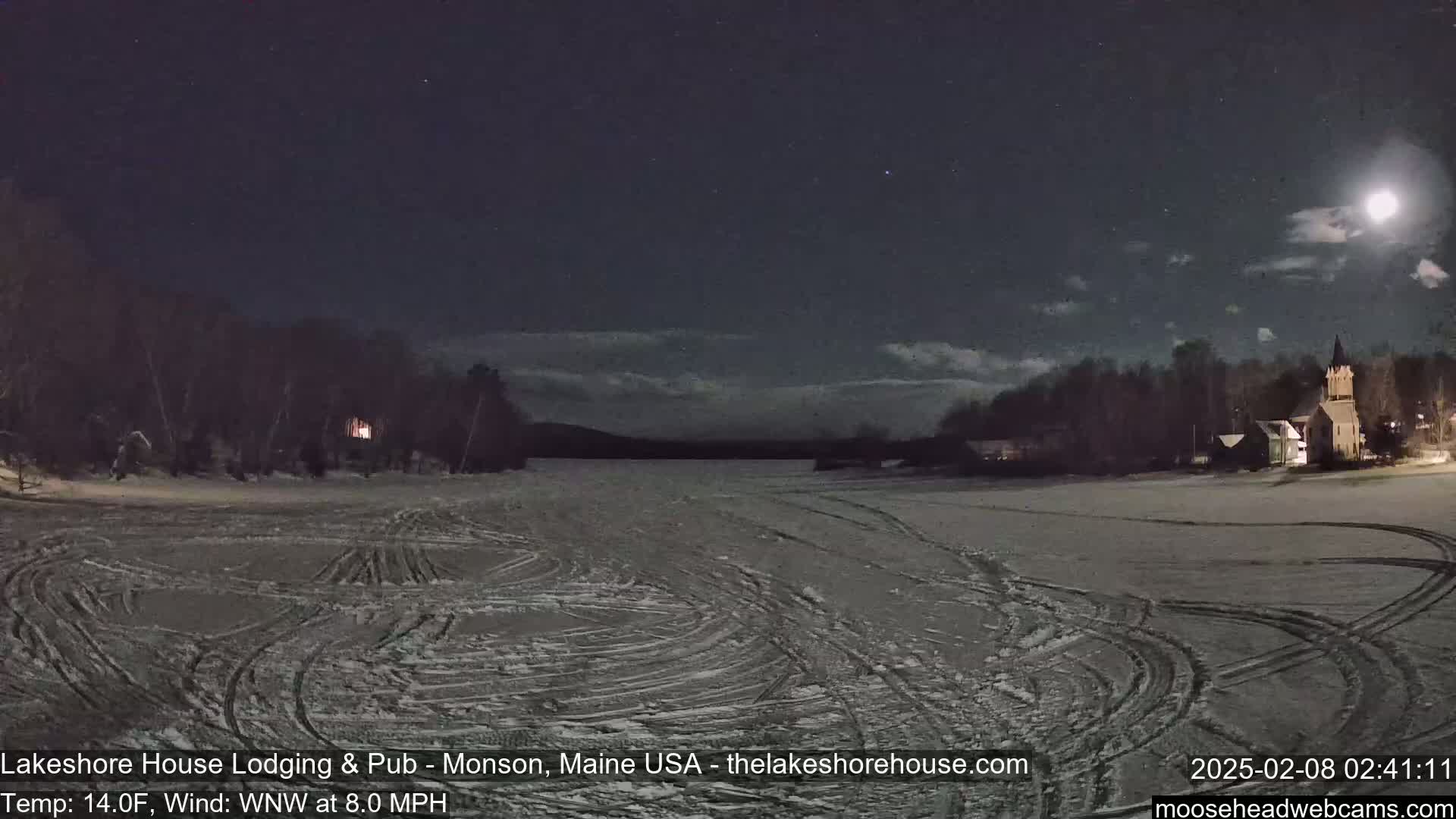 A snow-covered, frozen lake at night under a partly cloudy moonlit sky shows tire tracks and a building in the distance.