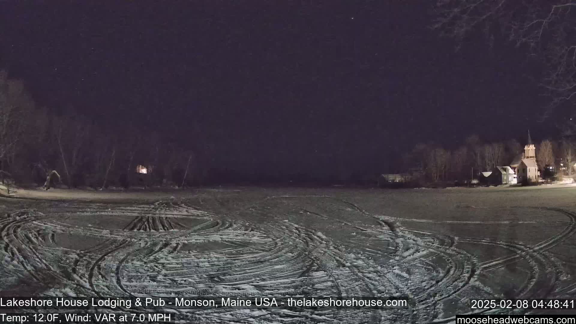 A snow-covered field with tire tracks under a dark, night sky shows a few buildings and trees in the distance.