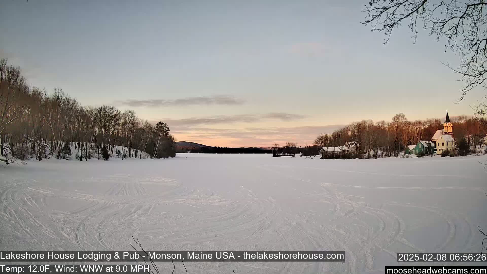 A frozen lake at sunset, with snow-covered trees and buildings lining the shore, under a pale pink and blue sky.