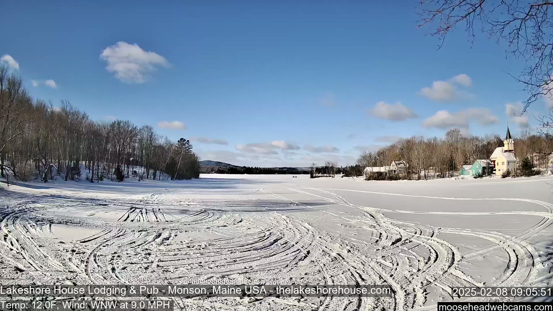 A frozen lake with tire tracks shows a clear, sunny day with a few clouds and snow-covered trees and buildings along the shoreline.