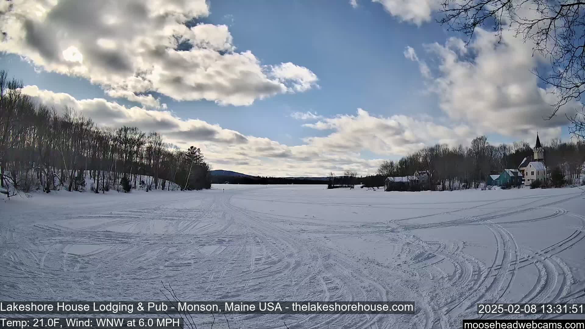 A snow-covered frozen lake with tire tracks, buildings on the far shore, and a partly cloudy sky.