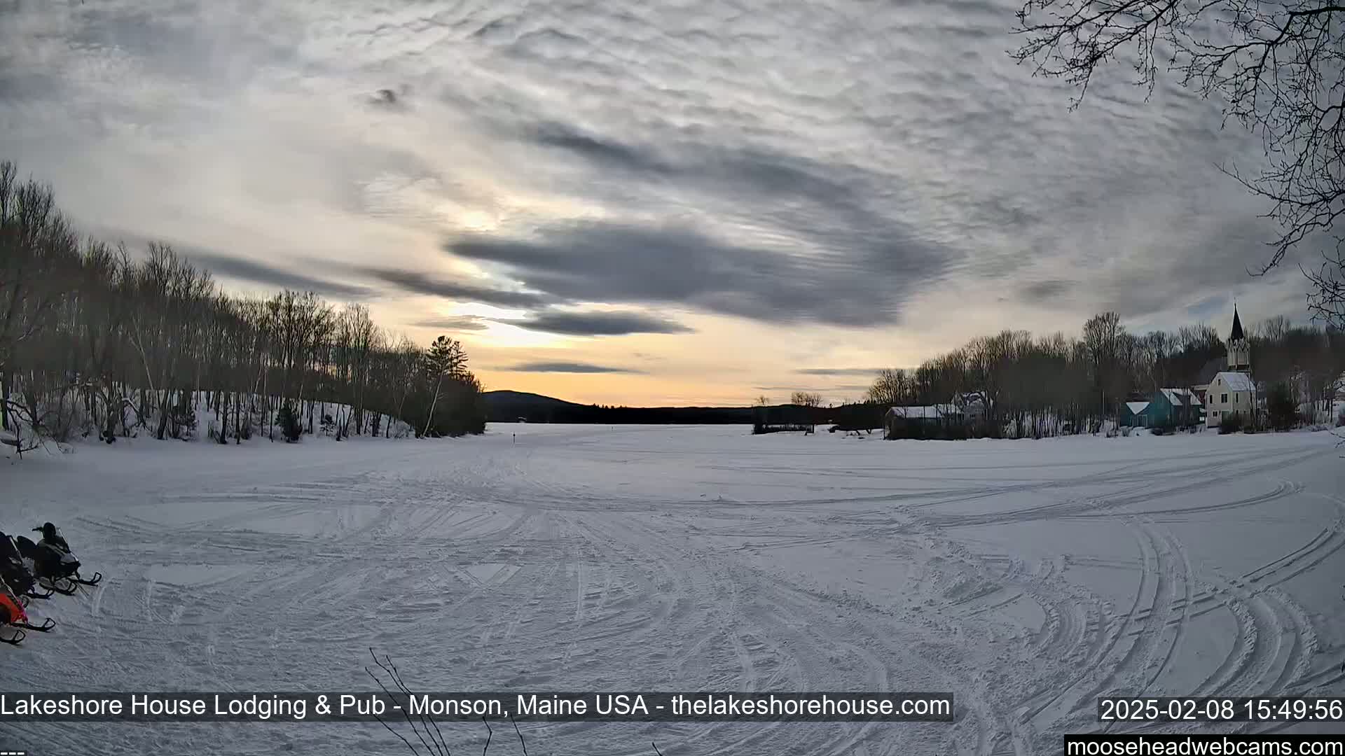Snowmobiles are parked on a frozen lake under a cloudy sunset, with a small town and trees visible in the distance.