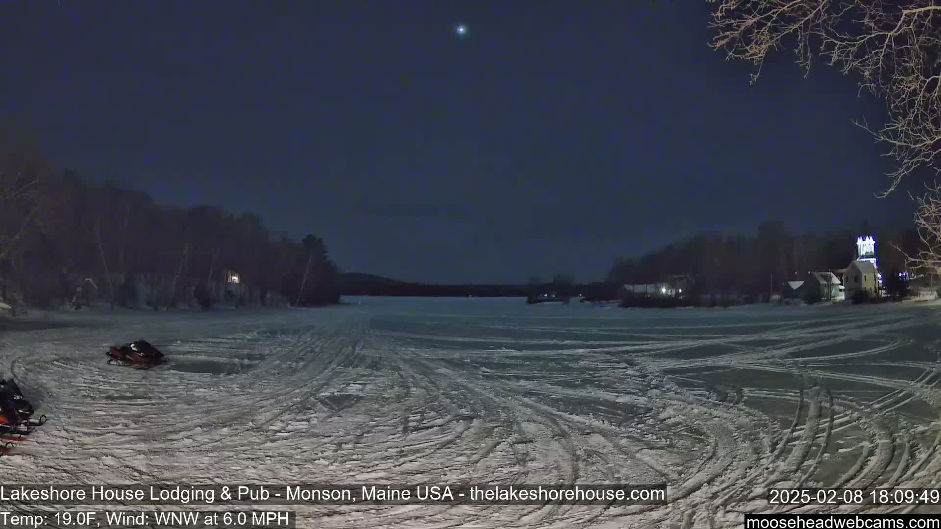 A snow-covered frozen lake at night, with snowmobiles parked on the ice, is viewed from a shoreline with buildings and trees visible in the distance under a dark, clear sky.