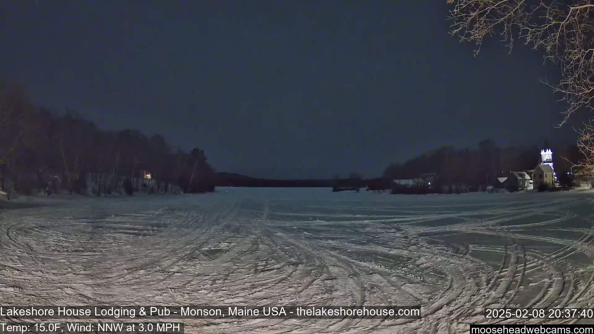 A snow-covered frozen lake is visible at night, under a dark sky, with trees and buildings along the shoreline.