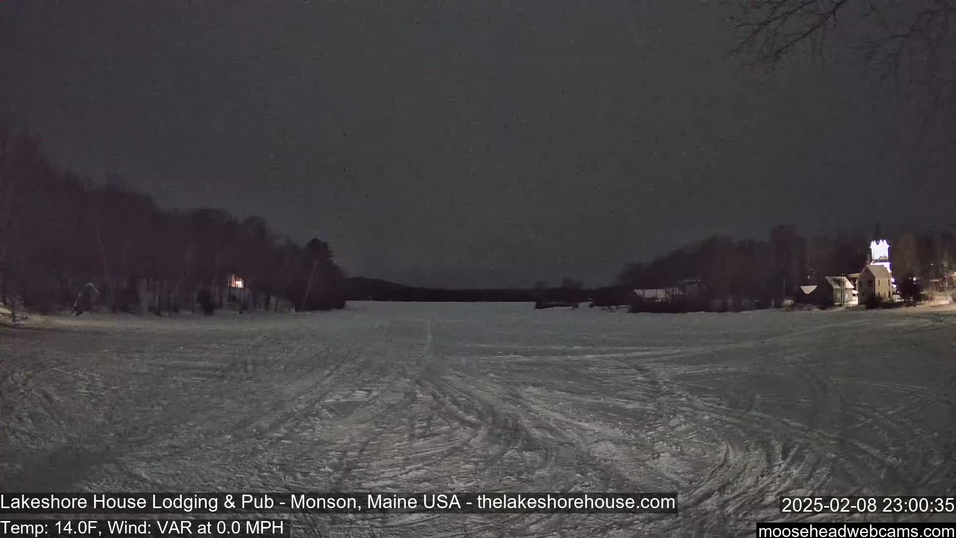 A frozen lake at night is visible, with a building lit up on the far right and dark trees along the shoreline under a dark sky.