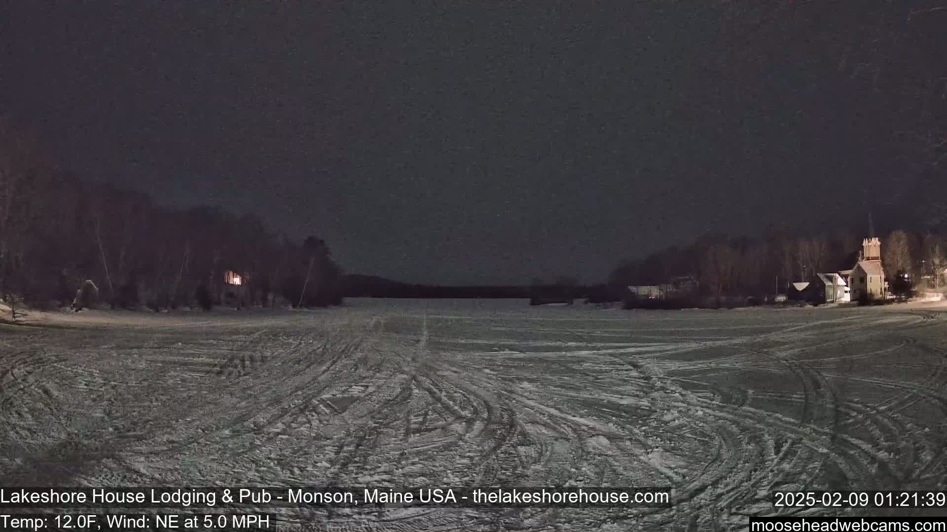 A snow-covered frozen lake at night, with a northeast wind and buildings visible in the distance under a dark sky.