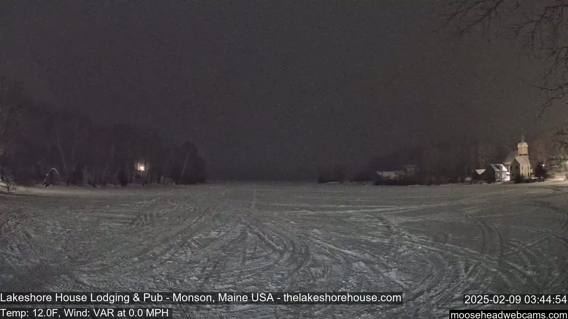 A snow-covered, dark field at night with sparsely lit buildings and trees in the background under a dark sky.