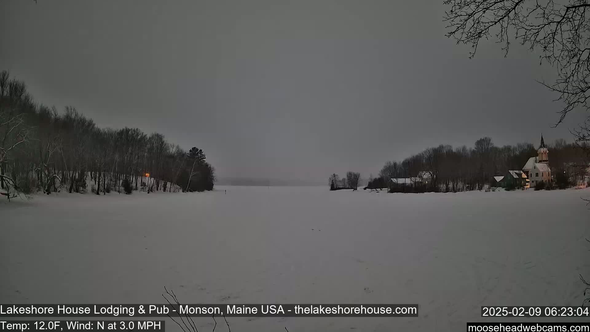 A snow-covered frozen lake is seen at dusk, with dark, snow-laden trees lining its shores and buildings visible in the distance.