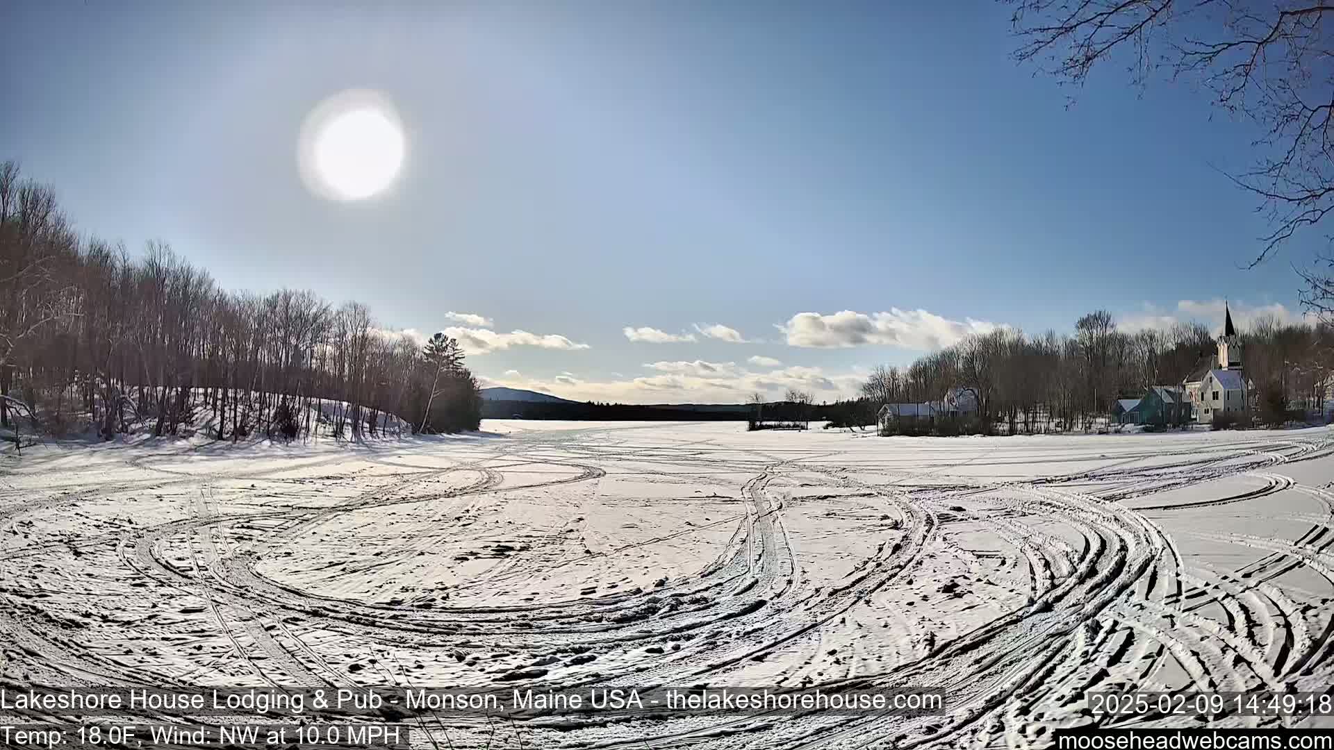 A snow-covered frozen lake with tire tracks shows a sunny, partly cloudy day, and a village with trees in the background.