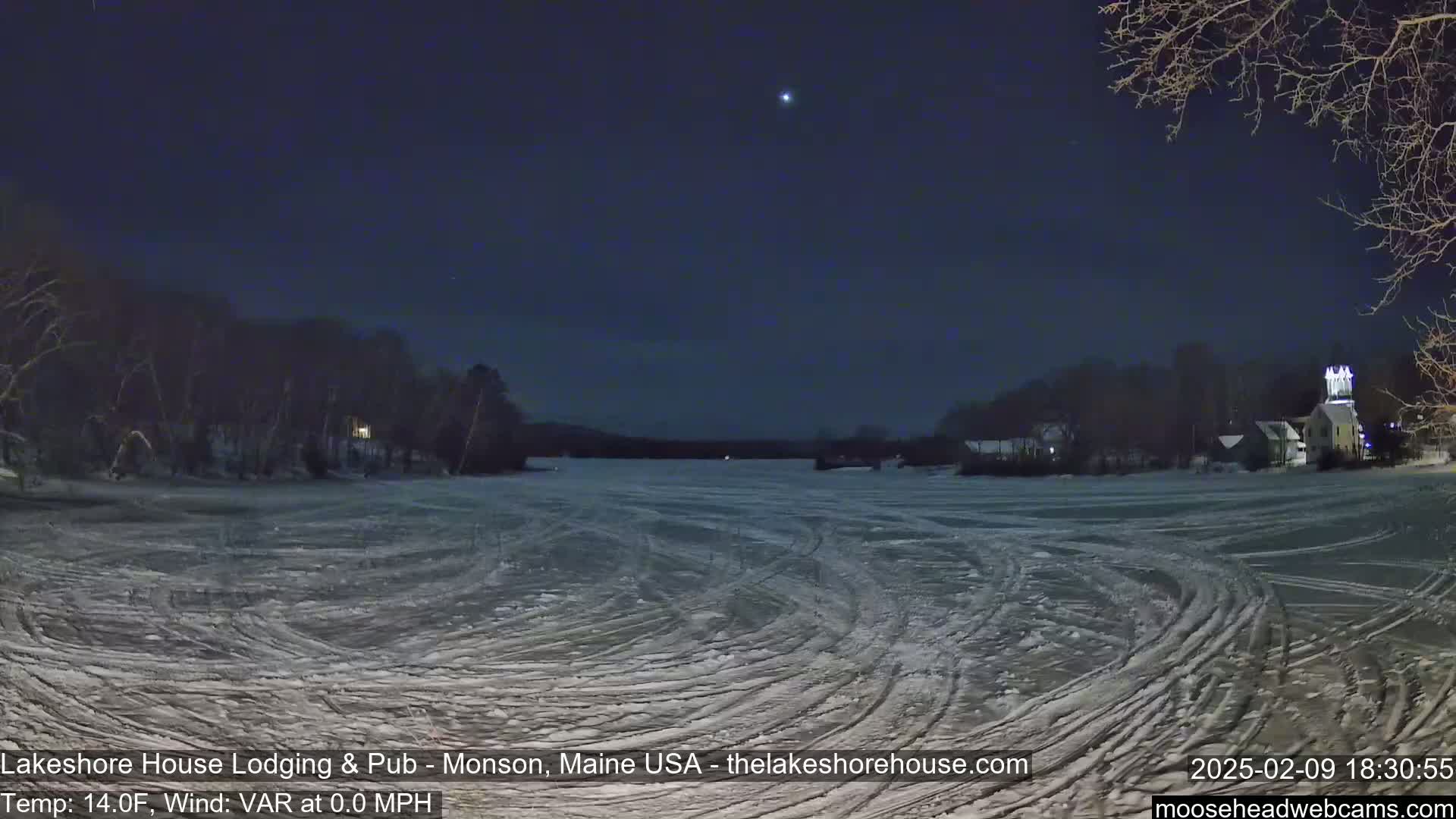 A frozen lake at night, under a clear, dark sky, shows tire tracks on the ice with snow-covered trees and buildings visible in the distance.