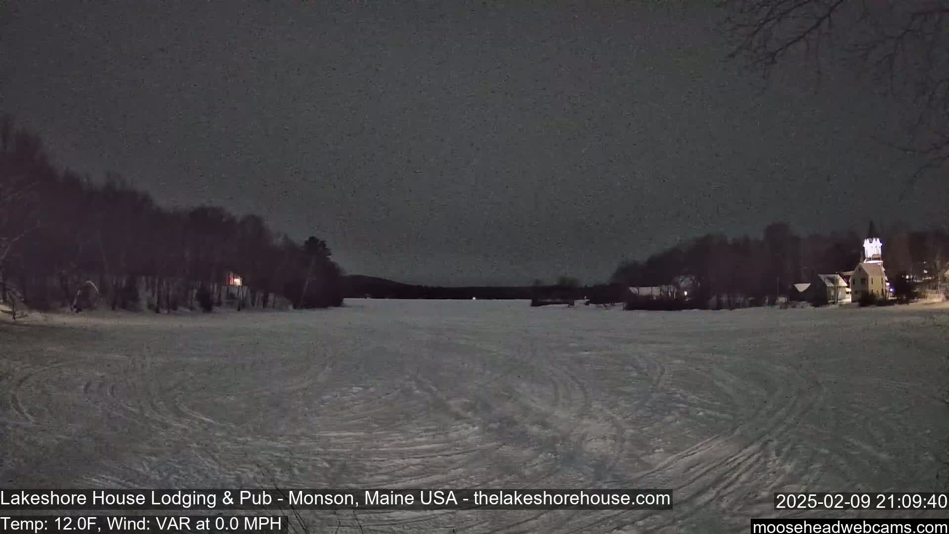 A snow-covered frozen lake at night is surrounded by dark trees and buildings with faint lights, under a dark, possibly snowy sky.
