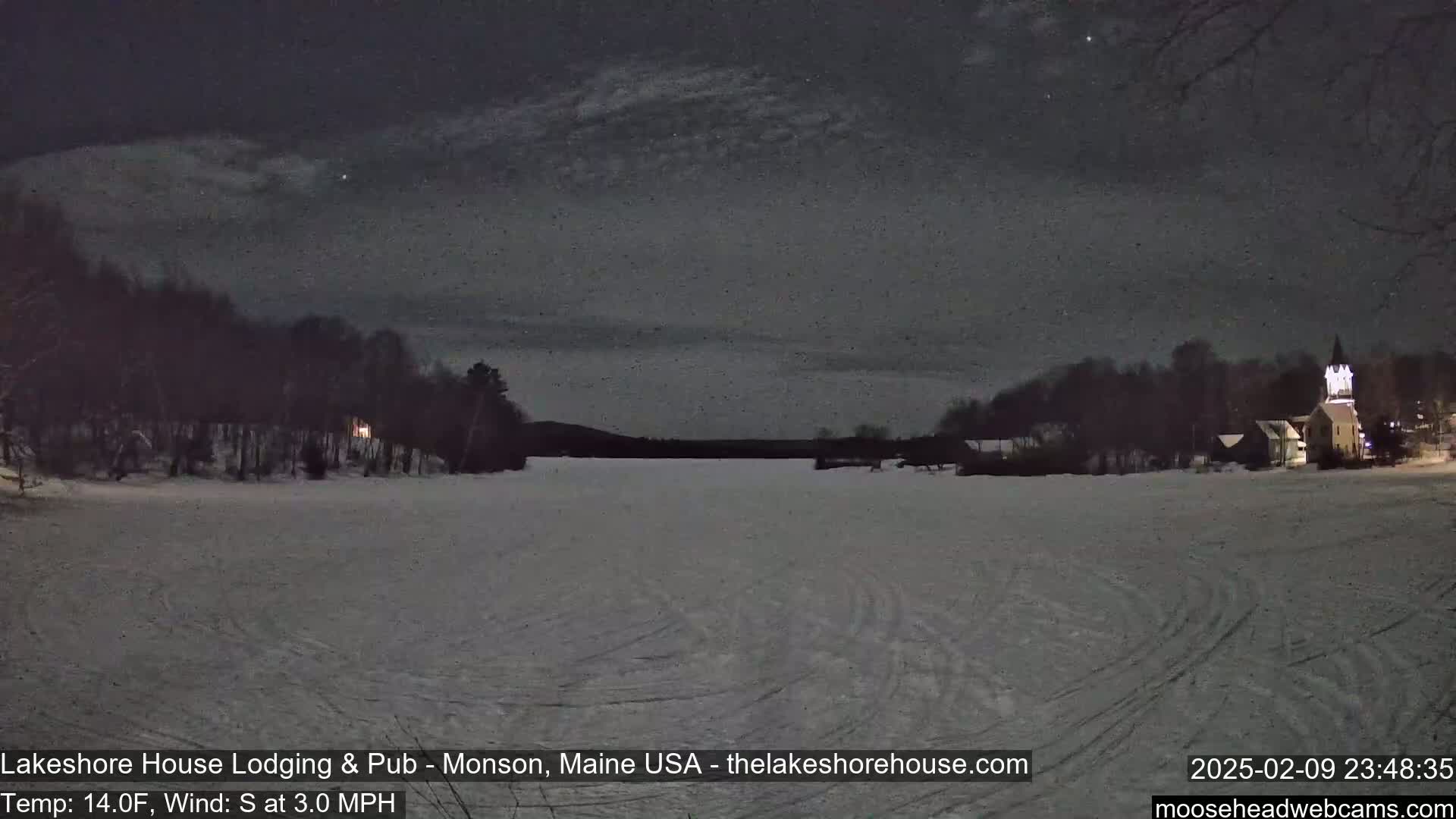 A snow-covered frozen lake is seen at night under a lightly clouded sky, with trees and buildings visible in the distance.