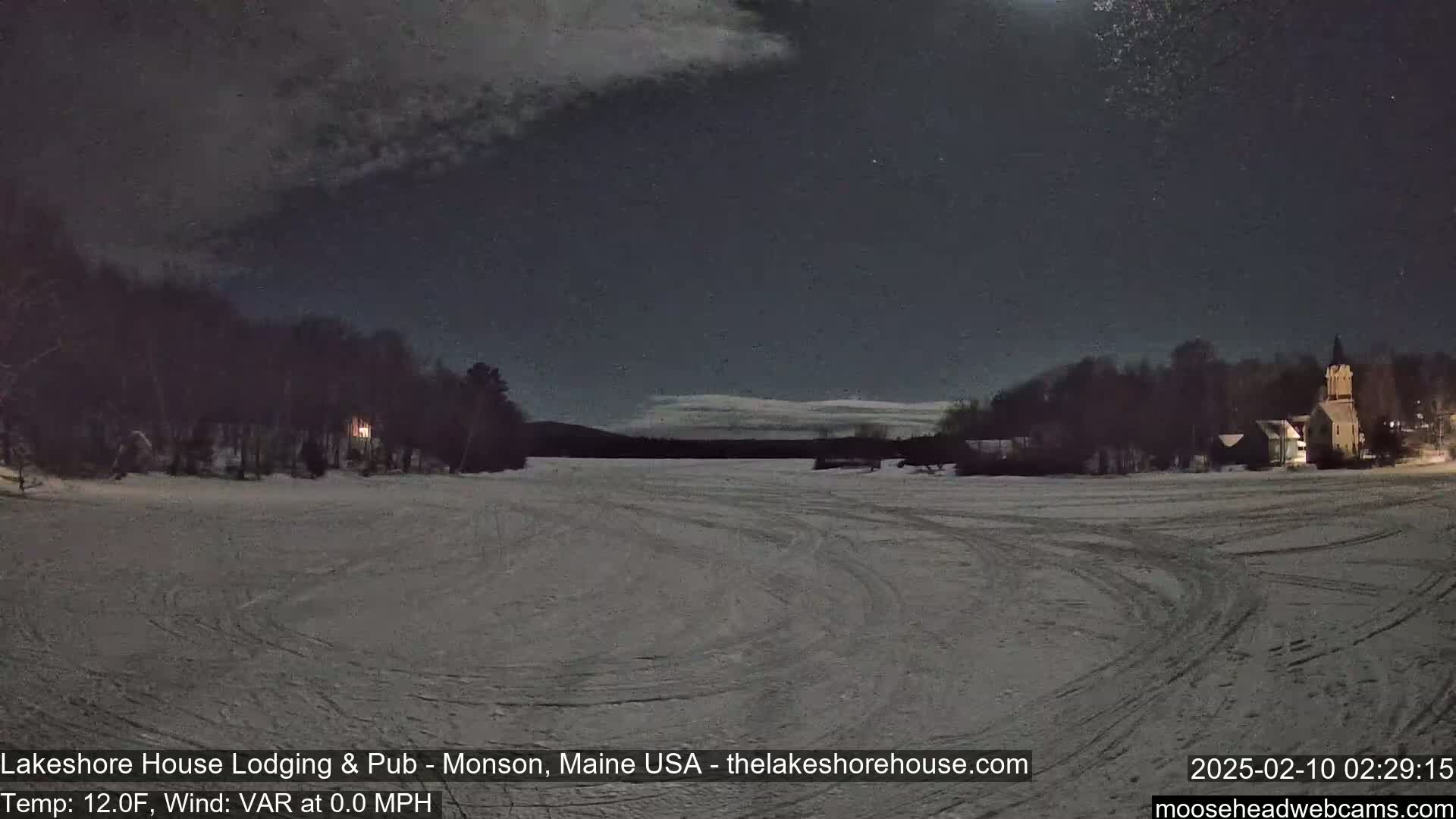 A snow-covered frozen lake is visible at night under a mostly clear sky, with buildings and trees along the shoreline faintly illuminated.
