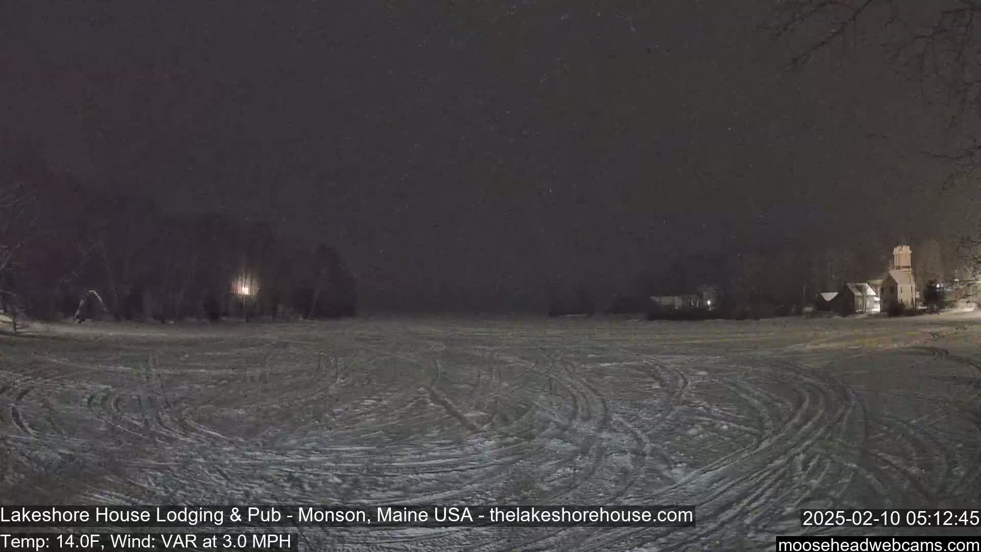 A snow-covered field with tire tracks is visible under a lightly snowing night sky, with dark trees and buildings in the background.