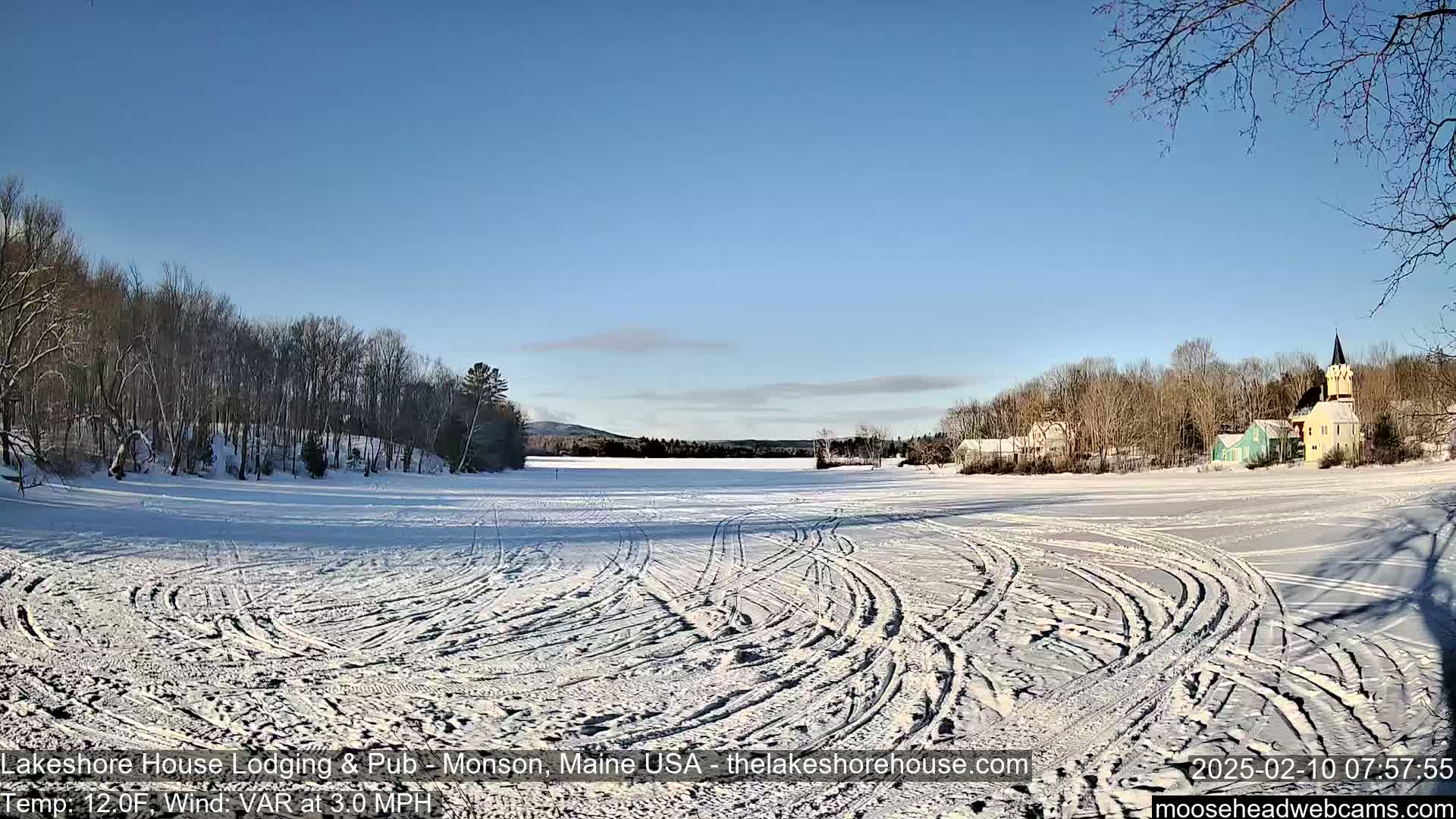 A snow-covered frozen lake with tire tracks and a village of houses and a church steeple in the background under a clear blue sky.