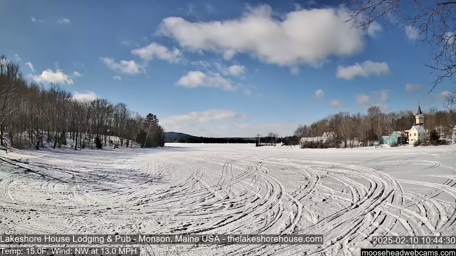 A snow-covered frozen lake with tire tracks shows a small town in the background under a partly cloudy, sunny sky.