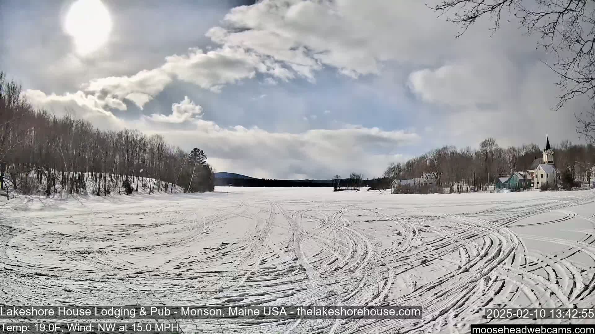 A snow-covered frozen lake with tire tracks, surrounded by bare trees and a village with a church steeple in the background, under a partly sunny sky with some clouds.