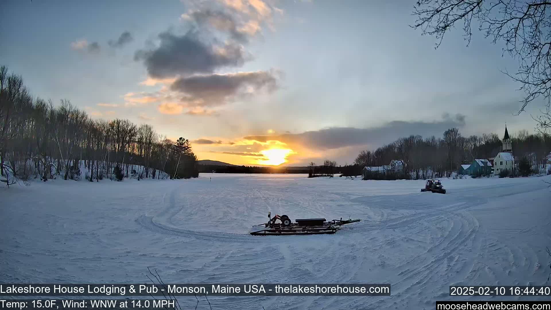 A snow-covered lake at sunset, with a small vehicle and buildings in the distance, under a partly cloudy sky and a 14 mph wind.