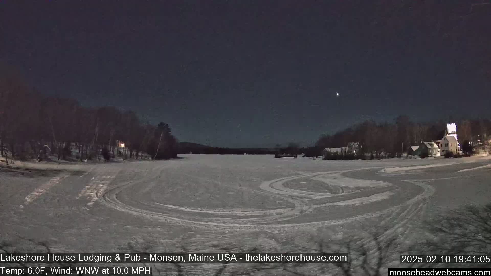 A frozen lake at night shows tire tracks on the ice, with buildings and trees visible along the far shore under a dark, clear sky.