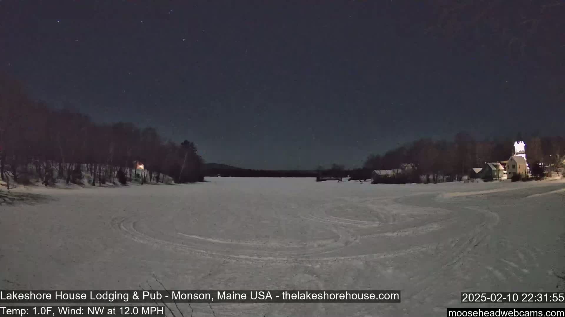 A snow-covered frozen lake is visible under a starry night sky, with a few buildings and trees lining the shore, and a northwest wind blowing at 12 mph with a temperature of 1°F.